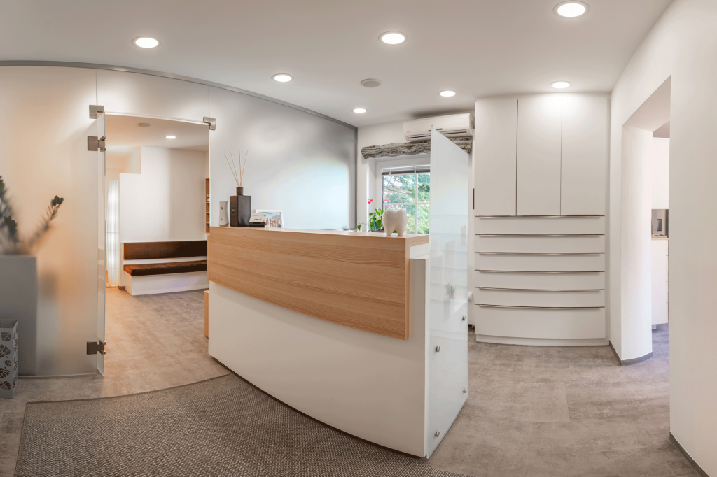 Heller und moderner Empfangsbereich der Zahnarztpraxis Krist in Kematen mit weißer Theke und Holzelementen.